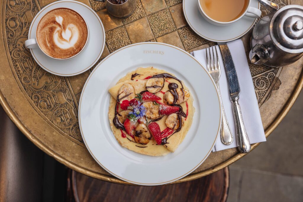 A table at a restaurant with a plate of pancakes topped with fruit and drizzled with chocolate sauce. A coffee and tea are also on the table, with cutlery to the side of the plate